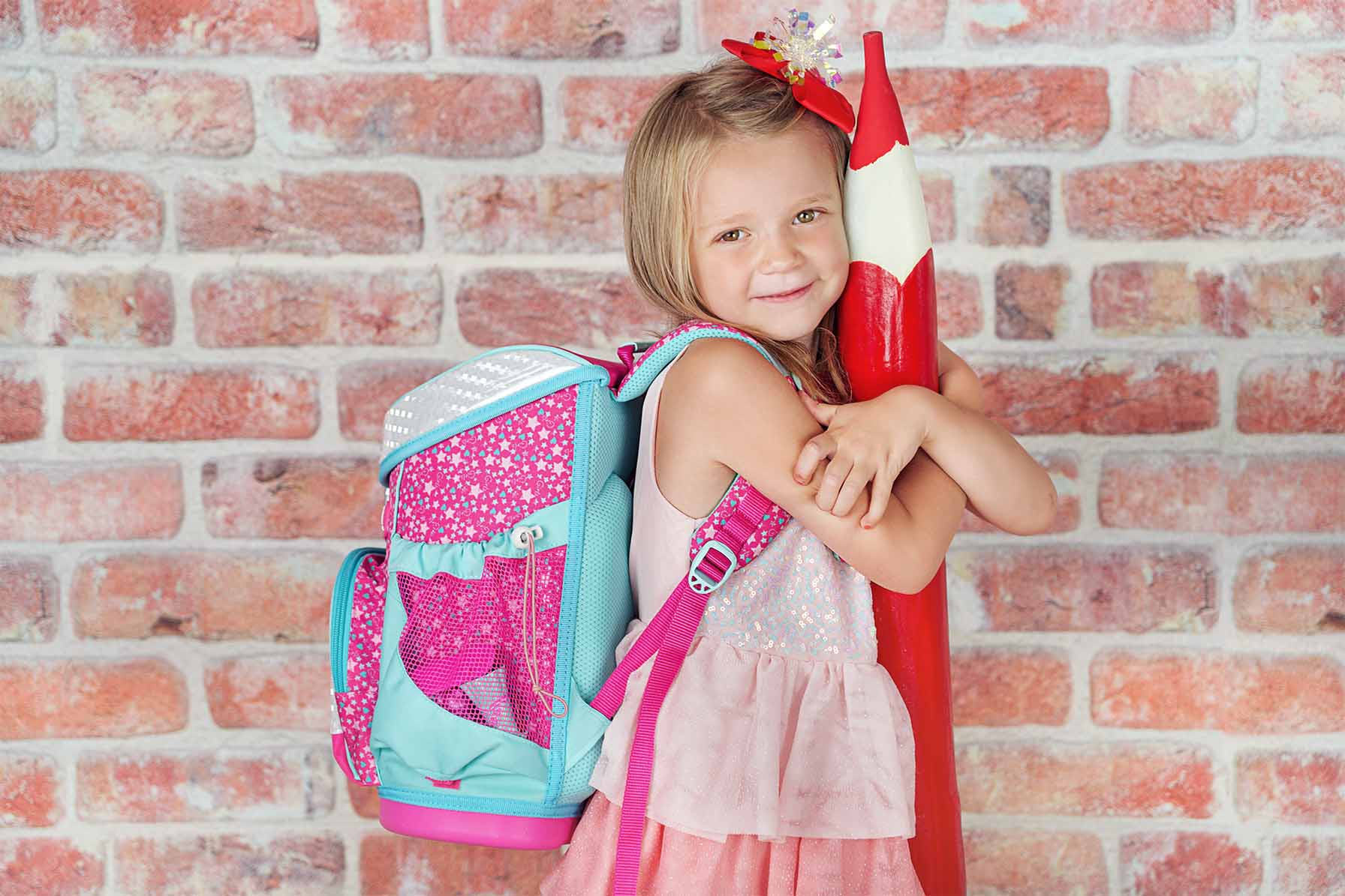 Little girl wearing a back-pack in front of her school smiles for a photo while holding a giant pencil.